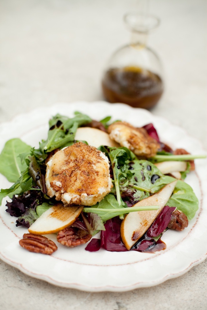 Greens with Warm Breaded Goat Cheese and Pears on white plate with small bottle of balsamic vinegar in background