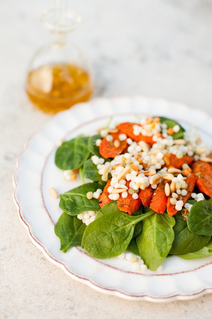 A white plate with a spinach and roasted carrot salad topped with couscous and a bttle of Easy White Balsamic Vinaigrette in the background