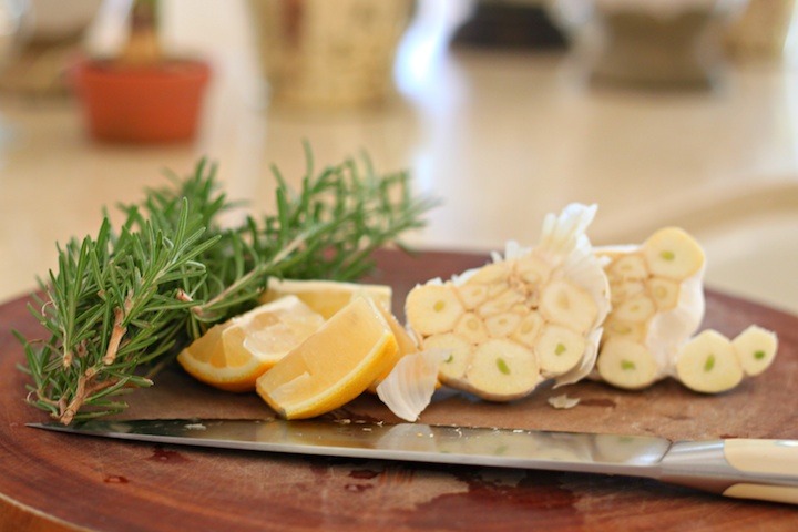 Two heads of garlic cut in half, three lemon wedges and sprigs of rosemary on a cutting board