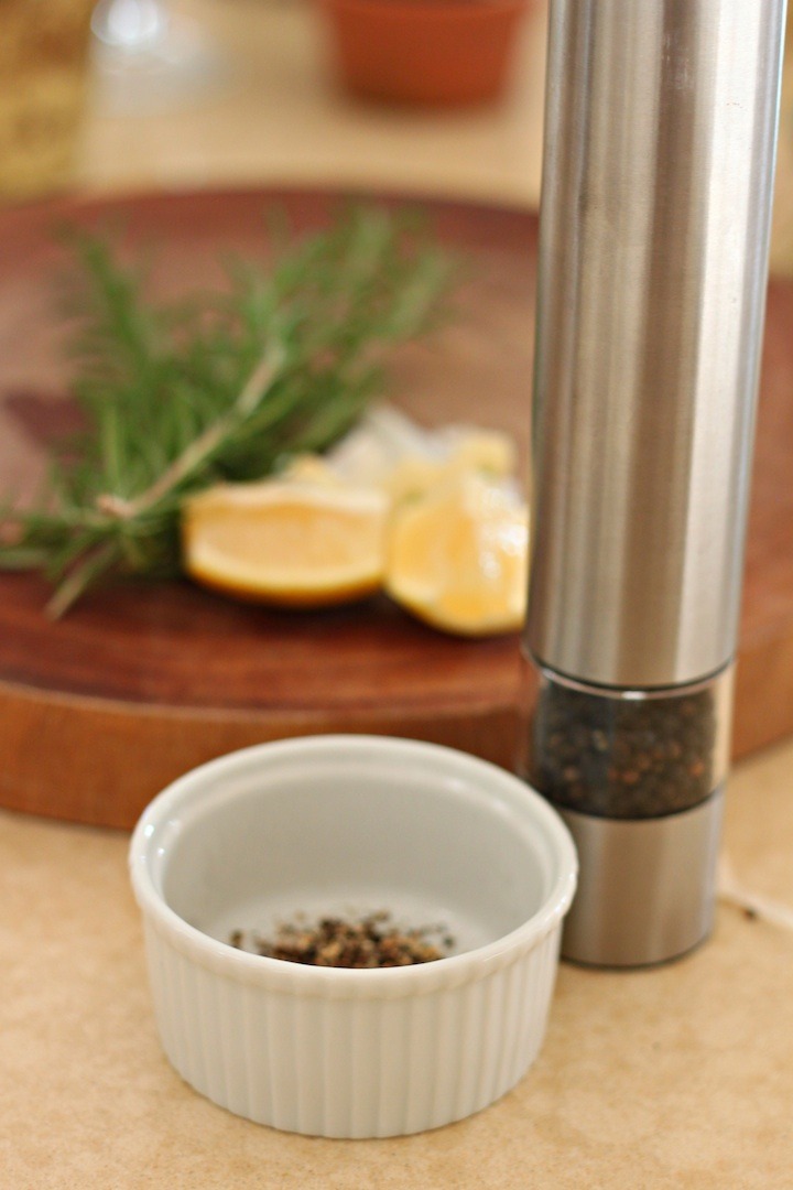A white ramekin and pepper grinder in front of a cutting board with lemon slices and rosemary springs to be used in Roasted Lemon/Garlic Chicken with Shallots