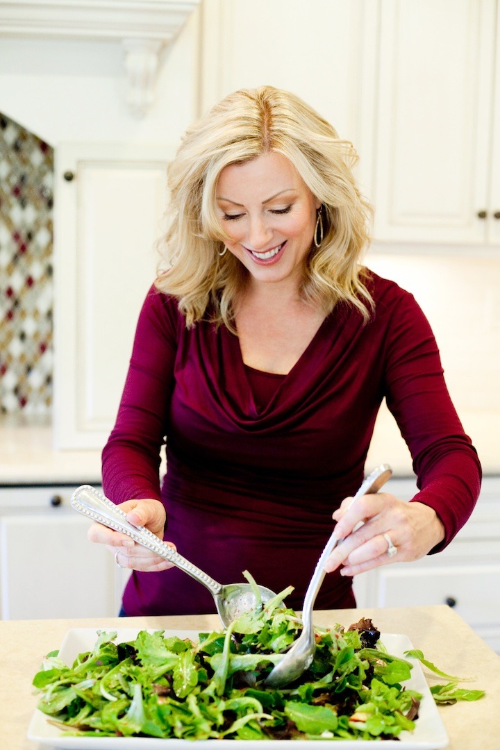 A blond woman in front of a counter using salad tongs to toss Greens with Pomegranate Vinaigrette