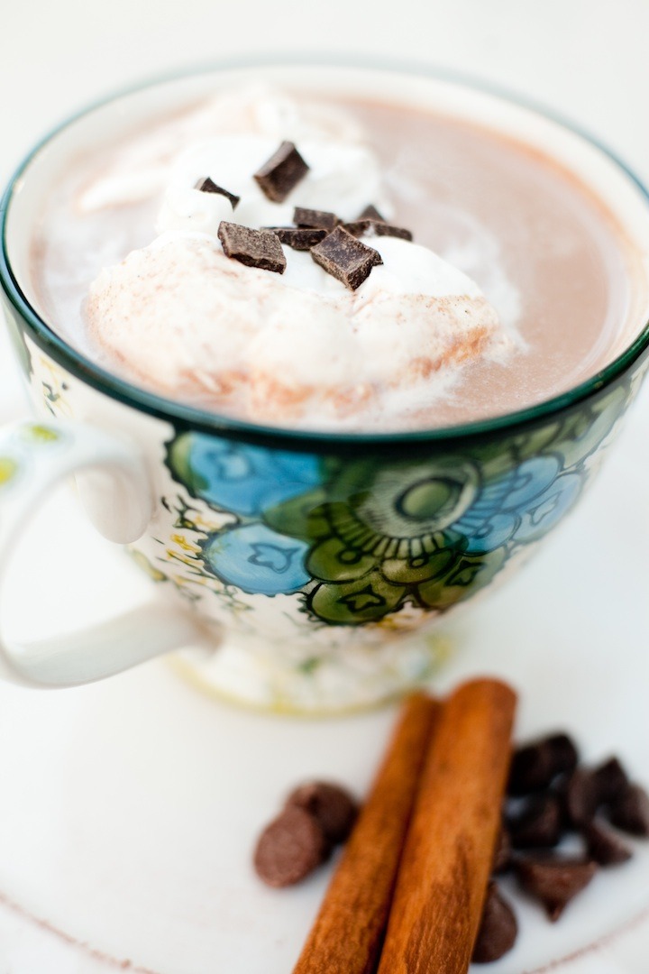 A colorful cup filled with hot chocolate and whipped cream with chocolate chips and cinnamon sticks in foreground