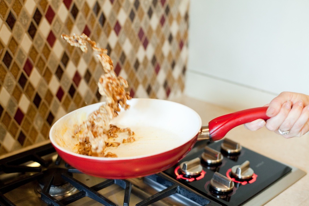 A woman's hand holding a red pan and flipping nuts in the air to demonstrate How To Caramelize Nuts