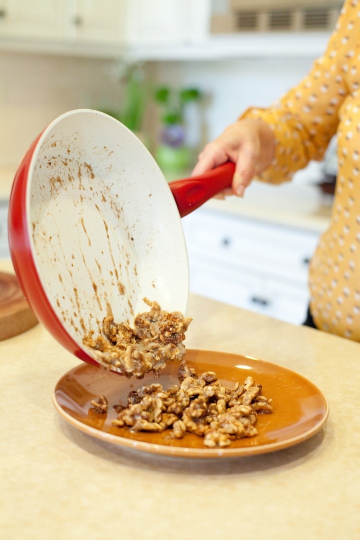 A woman's hand holding a pan and pouring caramelized nuts onto a plate to demonstrate How To Caramelize Nuts