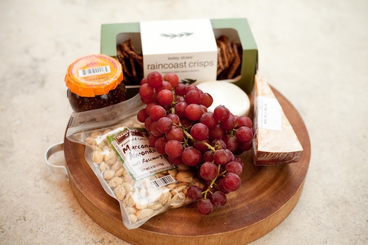 The making of the No fuss appetizer platter set on a cutting board: a jar of fig spread, marcona almonds ina bad, a wedge of gouda cheese, a round of goat cheese, a package of crackers, and one bunch of grapes