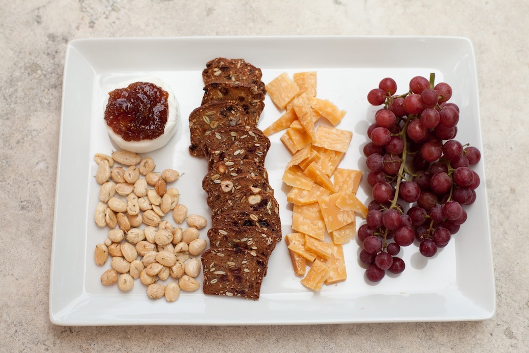 No fuss appetizer platter: A white platter with almonds. crackers, goat cheese, grapes, and gouda cheese, sitting on a marble surface