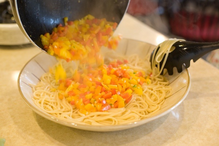 Sauteed peppers being poured into a large bowl of cooked linguini