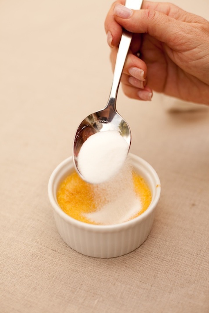 A female's hand using a spoon to sprinkle sugar in preparation to torch Lemon Vanilla Creme Brûlée