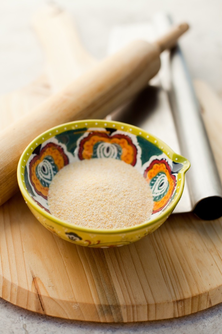 A wood cutting board with a small bowl of corn meal, a rolling pin and a pizza cutter; How to make Homemade Pizza Dough