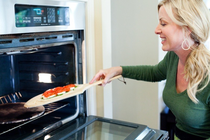 A blond woman using a pizza paddle to put pizza in the oven: How to make Homemade Pizza Dough 