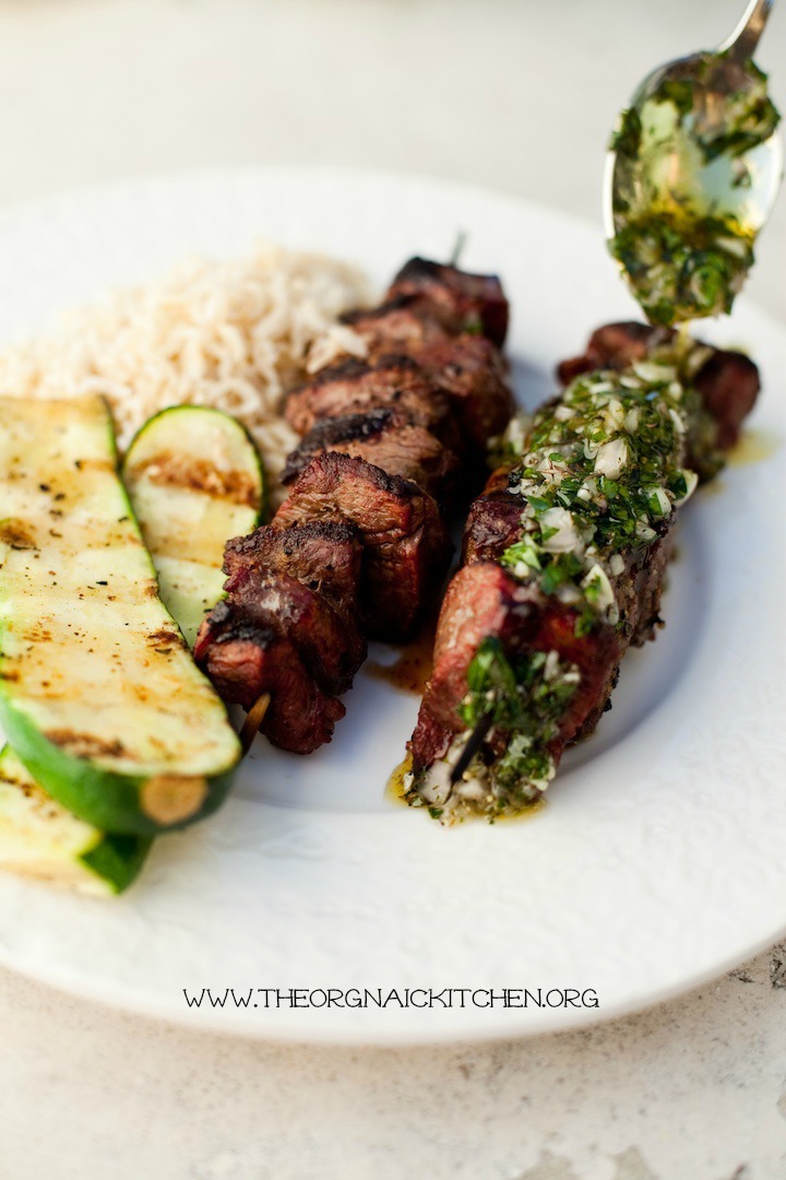 A white plate with steak kabobs, and two pieces of Simple Grilled Zucchini being drizzled with Chimichurri sauce