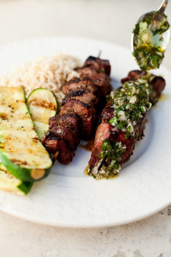 A white plate with Argentinian Beef Kabobs with Chimichurri Sauce and grilled zucchini