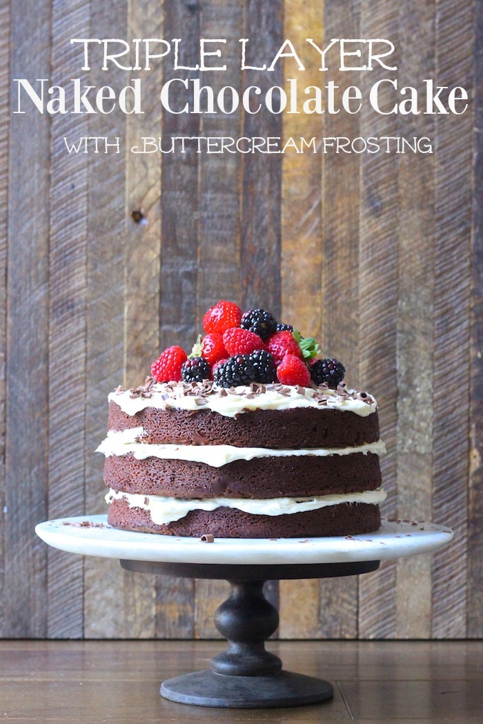 A triple layer Naked Chocolate Cake with Buttercream Frosting topped with berries on a cake plate in front of a wooden backdrop 