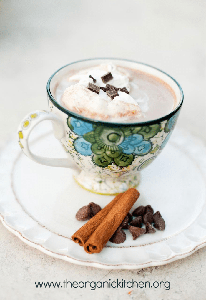 A mug of Spiced Hot Chocolate with Whipping Cream in a plate with chocolate chips and cinnamon sticks