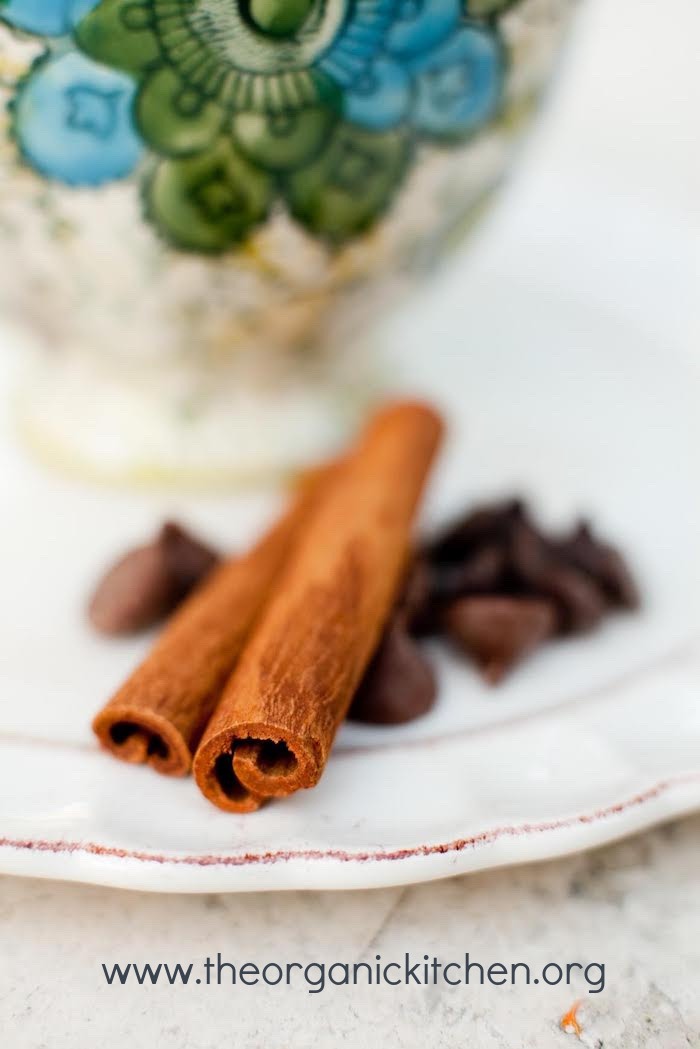 A close up of chocolate chips and cinnamon sticks on white plate with a mug of Spiced Hot Chocolate with Vanilla Whipped Cream