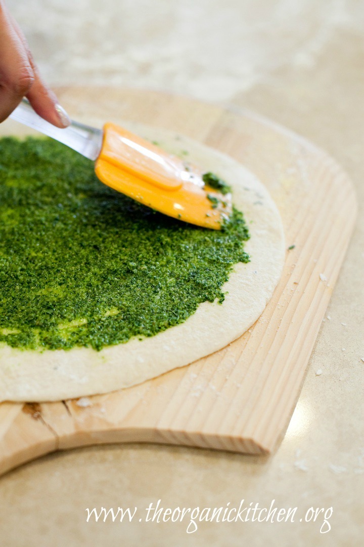 A woman's hand using a spatula to spread basil pesto on pizza dough: How to Make Basil Pesto from The Organic Kitchen