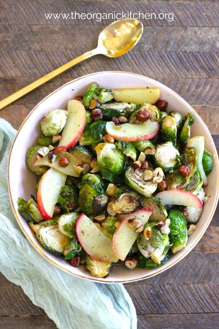 Roasted Brussels Sprouts with Apples and Hazelnuts in pink bowl on wood table with a gold spoon