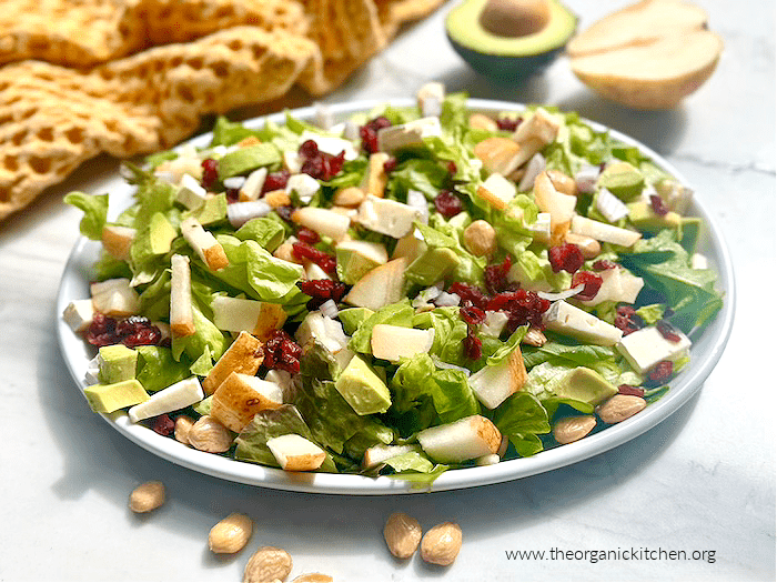Pear and Brie Salad with Honey Champagne Vinaigrette on white platter with almonds in the foreground and a half pear, half avocado and dish towel in the background