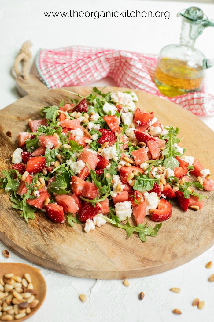 A large platter filled with Greens with Watermelon and Strawberries with a bottle of white balsamic vinaigrette in the background