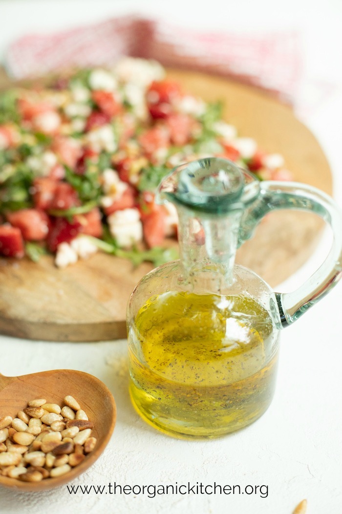 A bottle of White Balsamic Vinaigrette in front of a wood platter filled with Greens with Watermelon and Strawberries