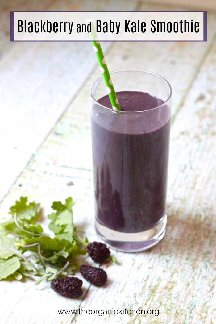 A tall glass of Blackberry and Baby Kale Breakfast Smoothie with green straw on a table with baby kale and blackberries scattered at the base of the glass