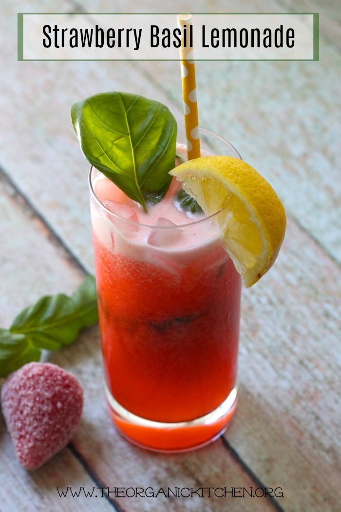 A tall glass of bright red Fresh Strawberry Basil Lemonade garnished with lemon wedges and basil leaves