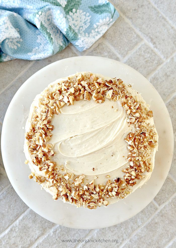 Hummingbird Cake (with gluten free option) garnished with walnuts on white cake plate with blue dish towel in the background