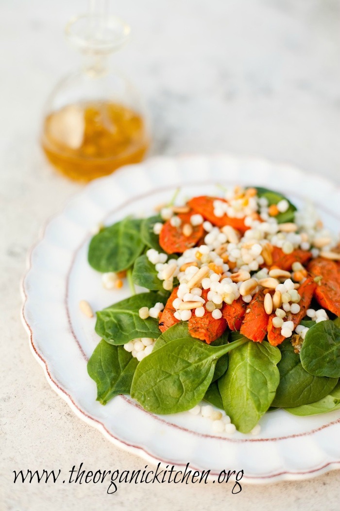 A small white plate with Baby Spinach Salad with Roasted Carrots and Israeli Couscous with small bottle of white balsamic vinaigrette in background