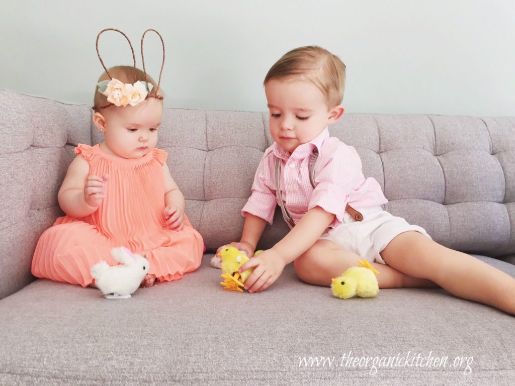 A baby girl and little boy on a sofa playing with toy baby chicken