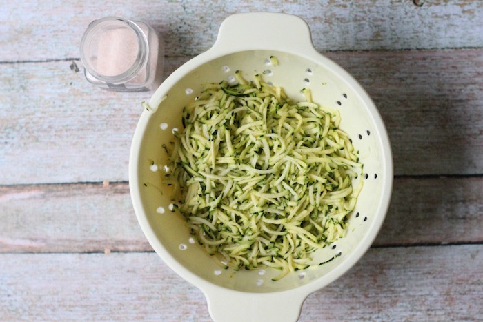 A colander full of grated zucchini with a jar of sea salt next to it
