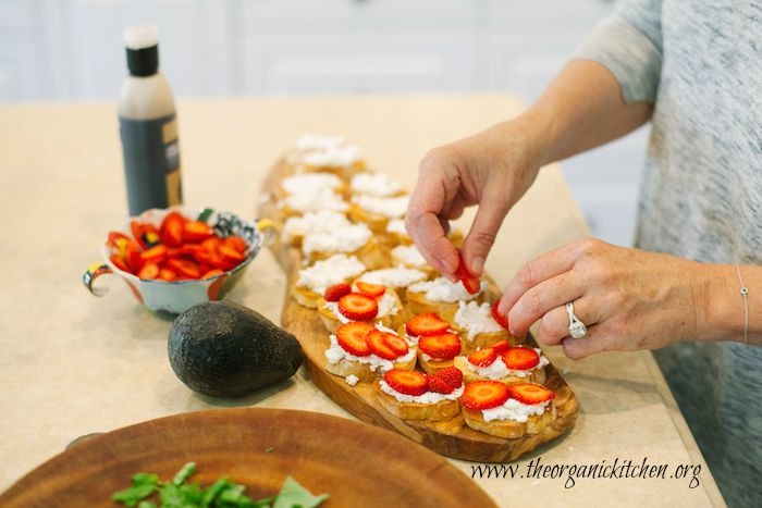 A woman placing strawberry slices on baguette slices topped with ricotta cheese to make Strawberry and Avocado Bruschetta: Bruschetta California Style!