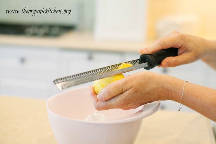Hands holding a microplane above a bowl to zest a lemon for Lemon Vanilla Raspberry Cake