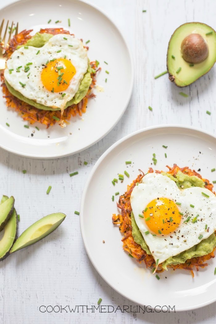 Sweet potato fritters topped with avocado mash, heart shaped fried eggs and chives on white plates, one of 12 Healthy, Delicious (Whole 30) Breakfast Recipes!