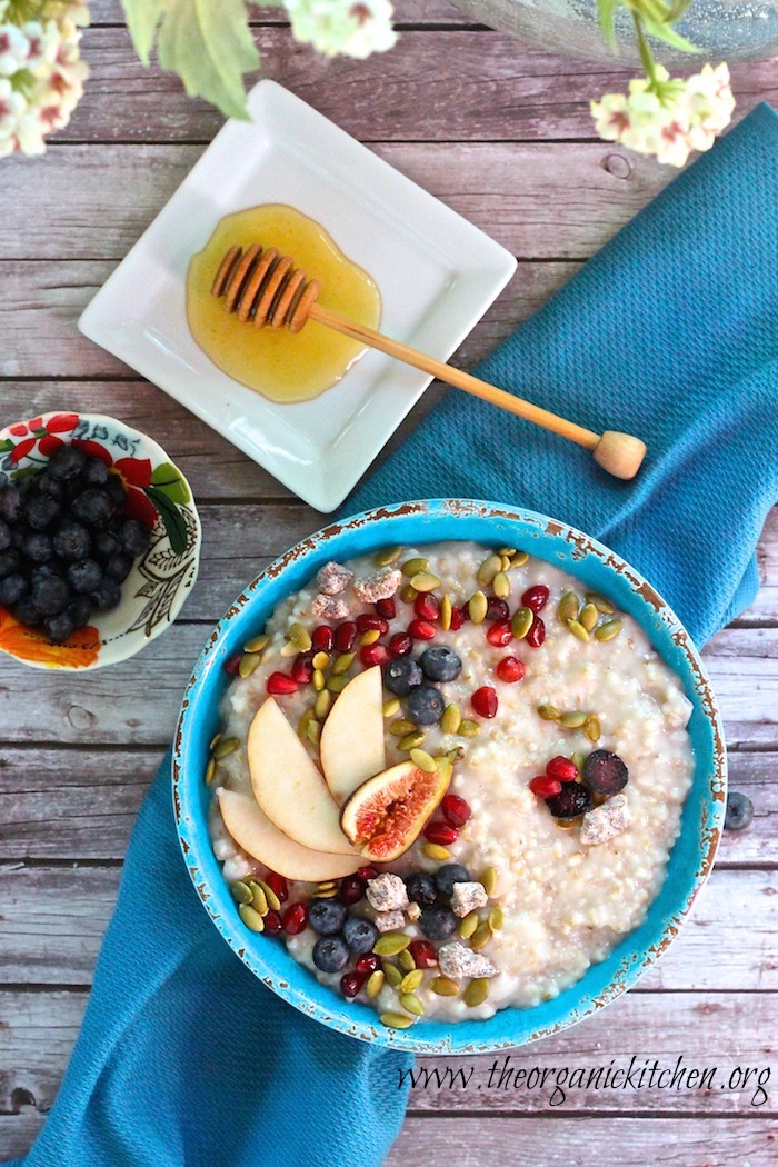 A blue bowl of Steel-Cut Oats with Seasonal Fruit on a table surrounded by flowers, berries and honey