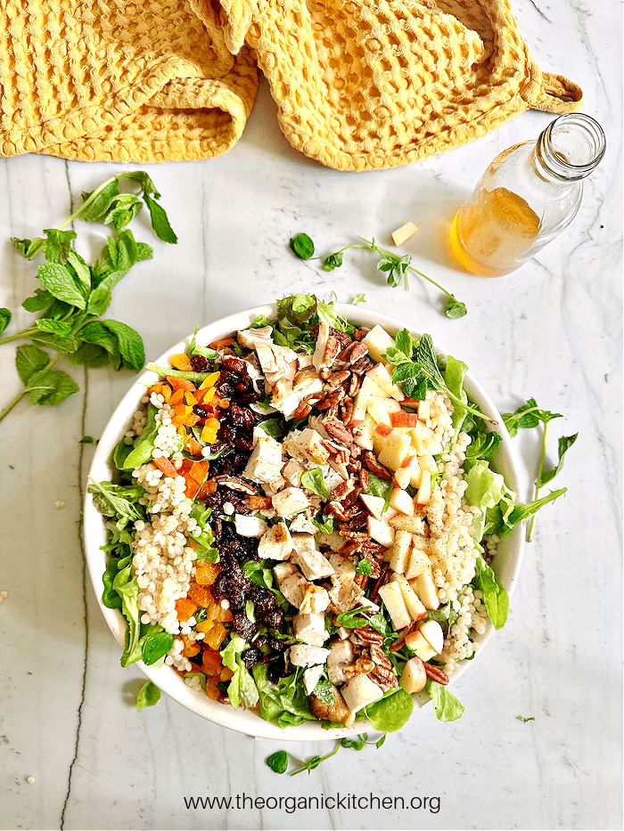 The Harvest Salad in a hwite bowl on marble counter with herbs, a bottle of dressing and a yellow dish towel above