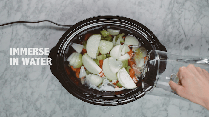 A cronk pot filled with vegetables with a woman's hand pouring water from a pitcher into the crock pot