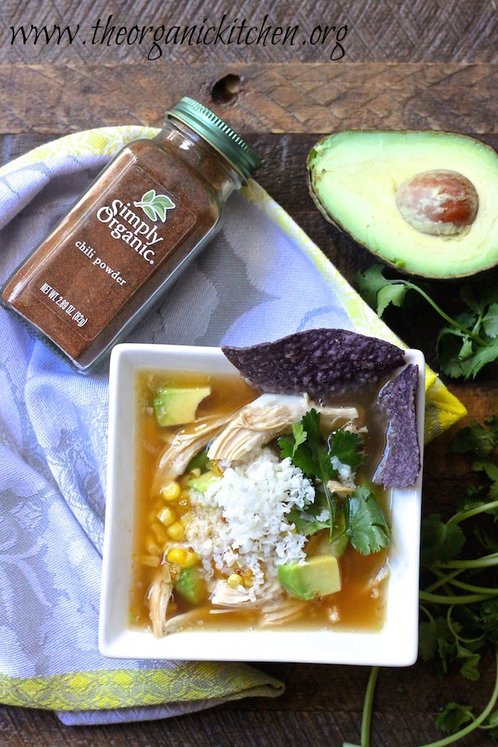 15 Minute Chicken Tortilla Soup in a square white bowl surrounded by avocado, herbs, and a bottle of chili powder