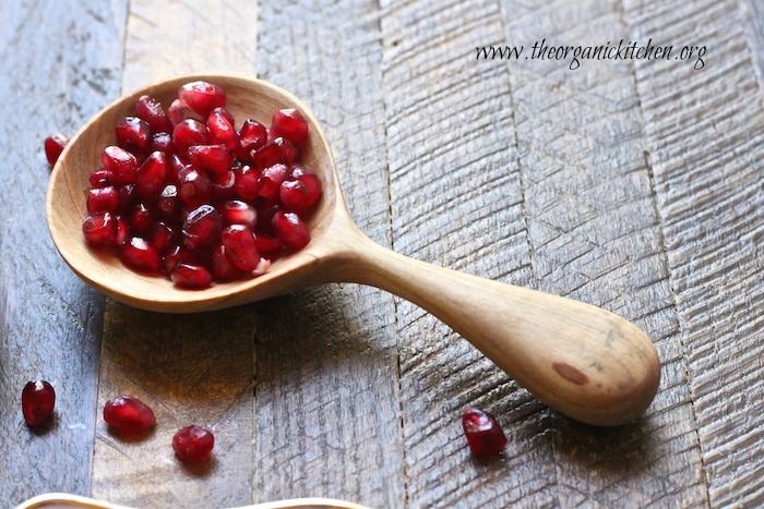 A wooden spoon filled with pomegranate seds set on a wooden cutting board