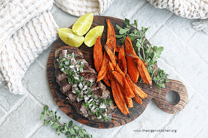 Steaks with Chimichurri and Roasted Harissa Sweet Potatoes on wood platter surrounded by lime wedges and oregano