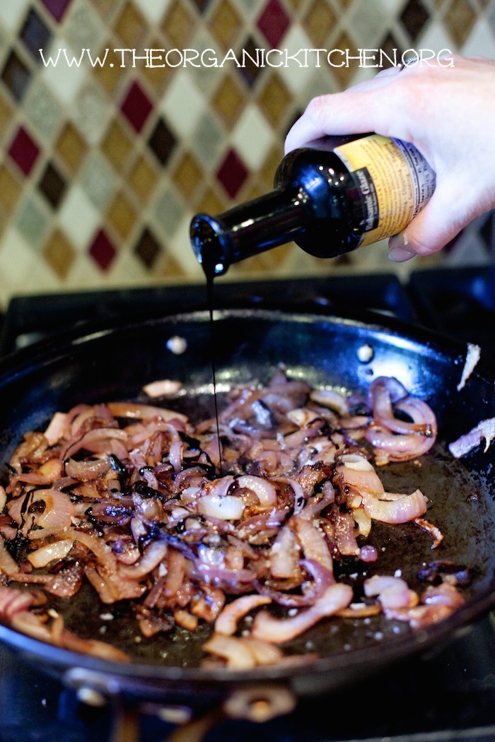 A pan on the stove filled with caramelized onions with a female's hand pouring balsamic glaze onto onions