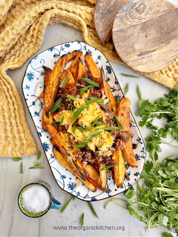 Sweet Potato-Black Bean Chili Cheese Fries on a blue and white dish with yellow dish towel, wooden serving spoons, and herbs in the background
