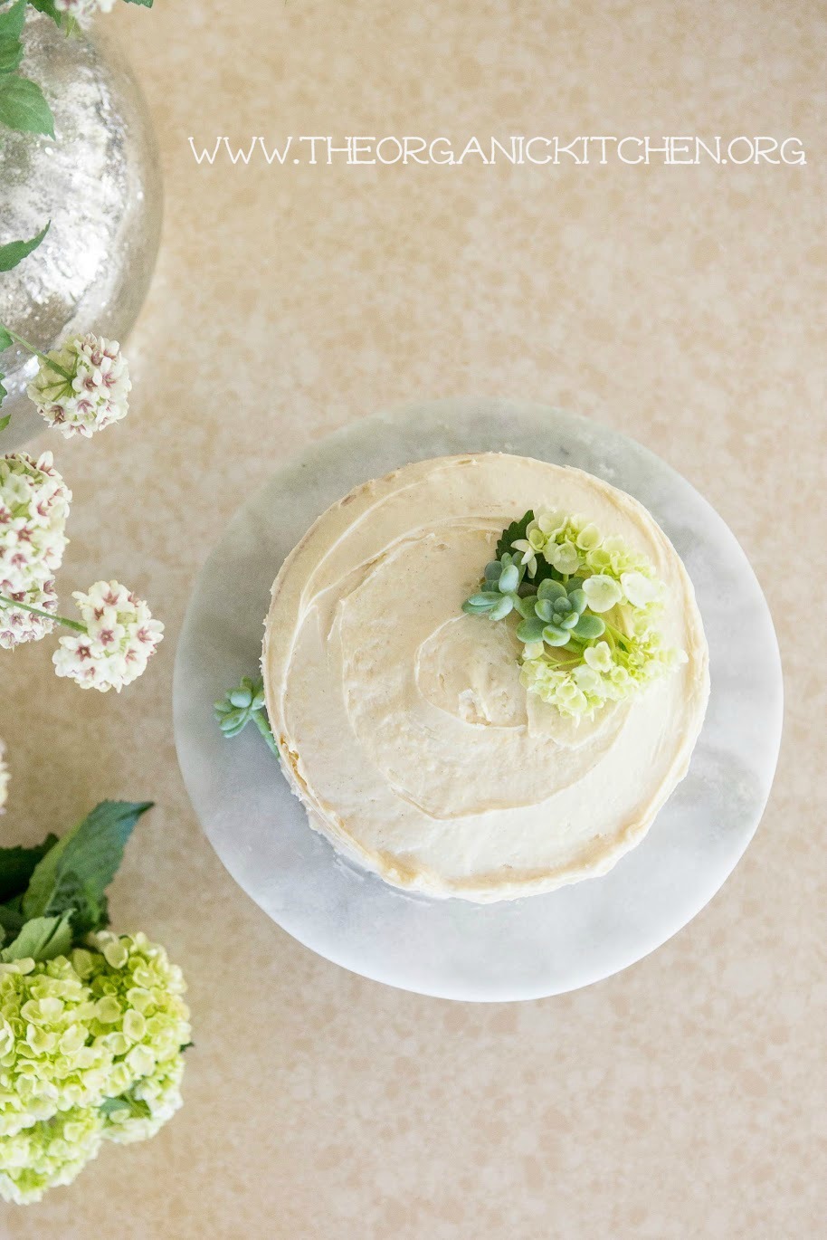 An aerial view of Buttermilk Cake with Vanilla Bean Cream Cheese Frosting set on a cake plate surrounded by flowers