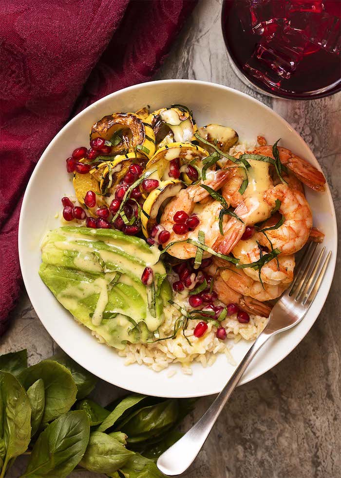 A white bowl roasted squash, avocado, shrimp and sauce garnished with pomegranate seeds on wood table as part of 12 Fantastic Rice Bowl Recipes!