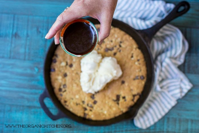 A woman's hand holding a small bowl of hot fudge ready to pour over an 'Almond Joy' Chocolate Chip Skillet Cookie! (Gluten free option)