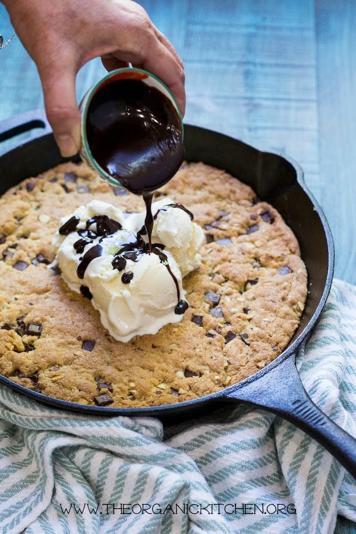 A woman's hand pouring a small bowl of hot fudge into an'Almond Joy' Chocolate Chip Skillet Cookie! (Gluten free option) topped with vanilla ice cream
