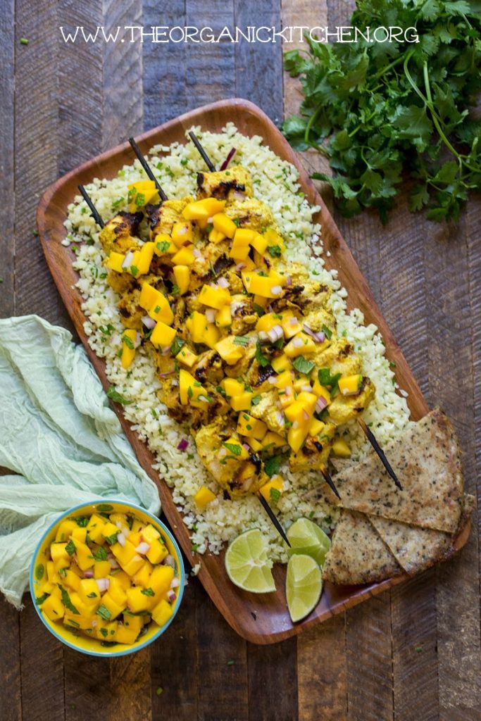 A wooden platter with Whole30 Tandoori Chicken skewers and a bowl mango Chutney on a brown wood backdrop 