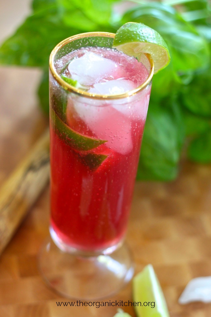 A tall glass of Pomegranate Lime Non-Alcoholic Spritzer on wood cutting board with basil in the background