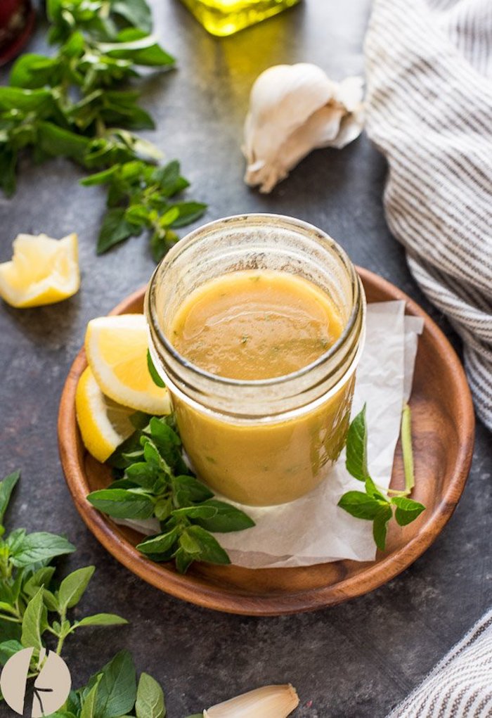 A jar filled with honey vinaigrette on a wooden plate surrounded by herbs and lemon wedges