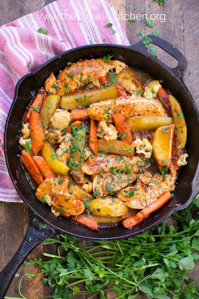 A red dish towel and parsley on a wooden table with a black cast iron skillet filled with One Pan Braised Chicken and Vegetables