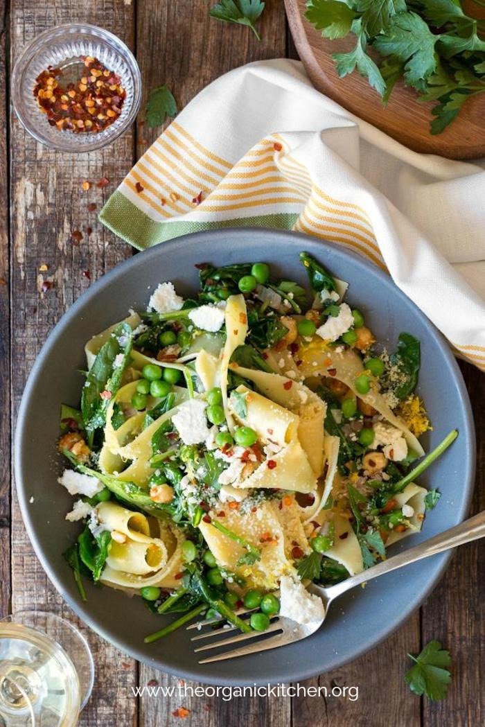A blue bowl filled with Pappardelle with Wilted Greens (Gluten Free Option) and surrounded by greens, pepper flakes and a napkin